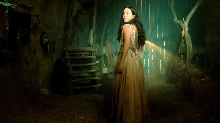 Schneewittchen, gespielt von Tijan Marei, steht in einem weißem Kleid in einem scheunenartigen Raum durch den ein wenig Licht durch das Tor fällt. Sie schaut neugierig über die Schulter.