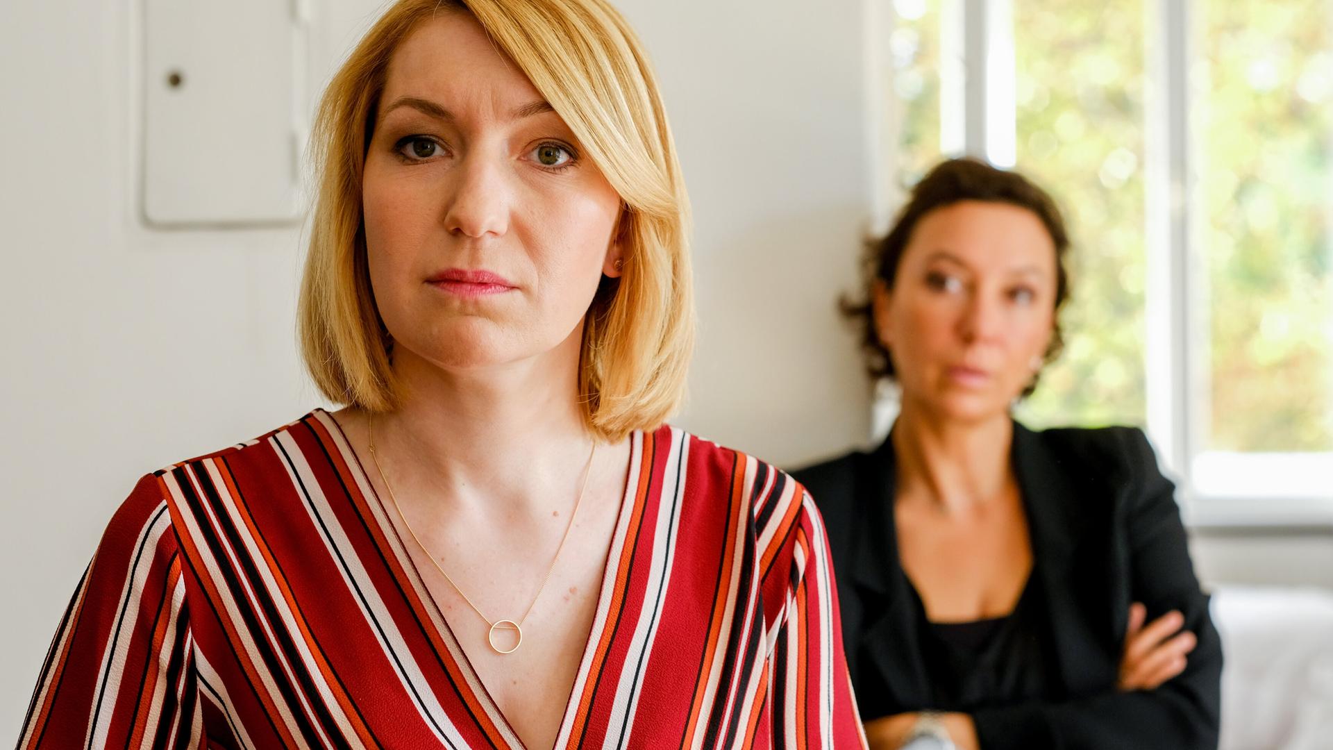 Eine blonde Frau blickt ernst in die Kamera, während im Hintergrund eine dunkelhaarige Frau mit verschränkten Armen sitzt und sie beobachtet. Die Szene spielt in einem hellen Raum.