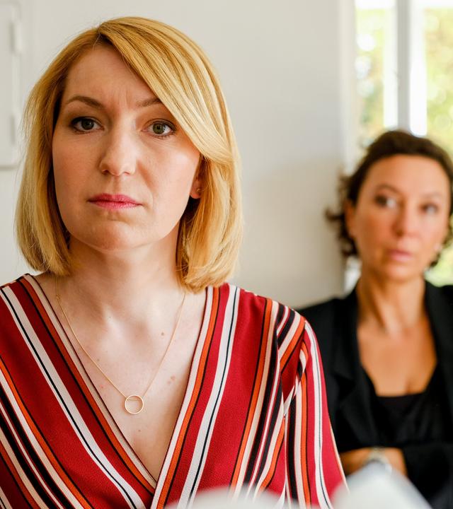 Eine blonde Frau blickt ernst in die Kamera, während im Hintergrund eine dunkelhaarige Frau mit verschränkten Armen sitzt und sie beobachtet. Die Szene spielt in einem hellen Raum.