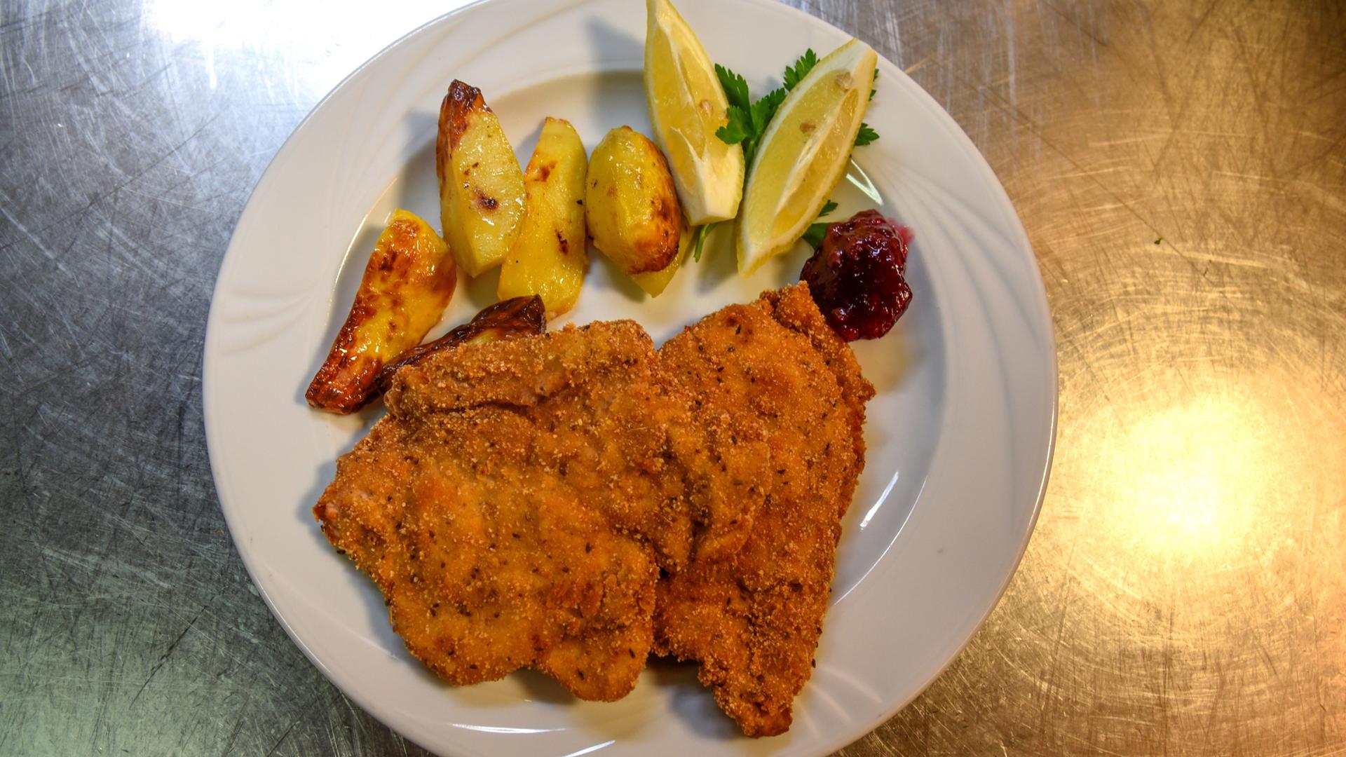 Das Bild zeigt einen Teller mit einem Cordon bleu, das mit einer Schüttelbrotpanade zubereitet ist. Das Cordon bleu ist gefüllt mit Speck und Ziegenkäse. Auf dem Teller befinden sich außerdem einige Stücke gebratene Kartoffeln in unterschiedlichen Formen, die goldbraun angeröstet sind. Dazu sind zwei Zitronenschnitze sowie ein Klecks Preiselbeermarmelade dekorativ angerichtet. Der Teller ist weiß und steht auf einer glänzenden, metallischen Unterlage, die silberfarben aussieht.
