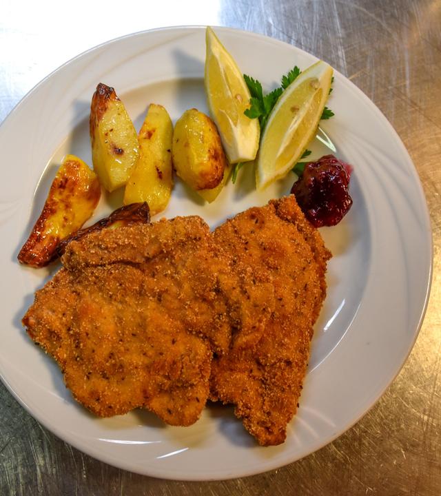 Das Bild zeigt einen Teller mit einem Cordon bleu, das mit einer Schüttelbrotpanade zubereitet ist. Das Cordon bleu ist gefüllt mit Speck und Ziegenkäse. Auf dem Teller befinden sich außerdem einige Stücke gebratene Kartoffeln in unterschiedlichen Formen, die goldbraun angeröstet sind. Dazu sind zwei Zitronenschnitze sowie ein Klecks Preiselbeermarmelade dekorativ angerichtet. Der Teller ist weiß und steht auf einer glänzenden, metallischen Unterlage, die silberfarben aussieht.