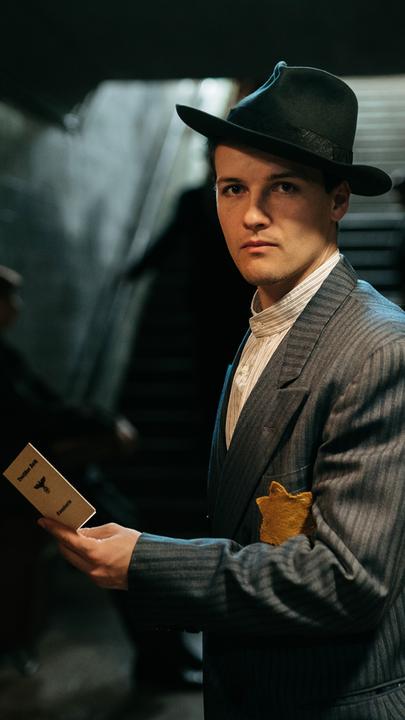 Spielszene: Cioma Schönhaus (Juri Senft) steht vor einer Treppe und hält seinen Ausweis in der Hand.