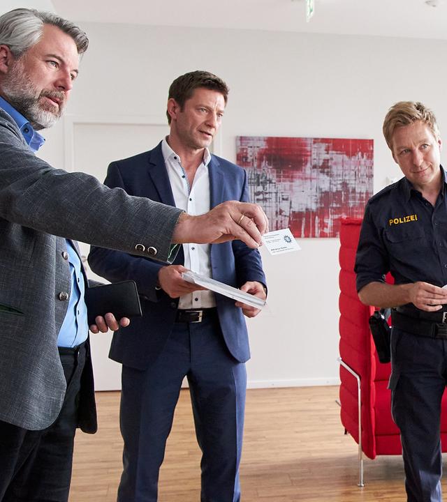 Die Kommissare Stadler (Dieter Fischer) und Hansen (Igor Jeftić) sowie Polizeihauptmeister Mohr stehen Maren Köhler (Lisa Fertner) in einem großen, mit Parkett ausgelegten Raum gegenüber und reden. Im Hintergrund sind diverse Sitzmöbel zu sehen sowie ein Bild an der Wand. Kommissar Stadler hält Maren Köhler seine Visitenkarte hin.