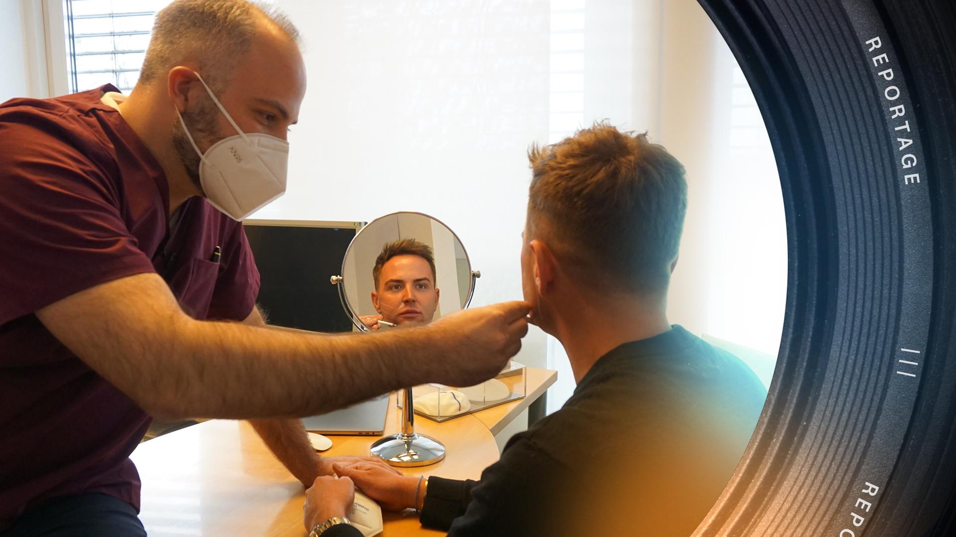 Zwei Personen sind in einem Raum zu sehen. Rechts sitzt ein Mann an einem Tisch, mit dem Rücken zur Kamera. Er blickt in einen Tischspiegel, sein Gesicht ist im Spiegelbild erkennbar. Links von ihm sitzt ein Mann mit Gesichtsmaske. Mit der rechten Hand hält er einen weißen Stift an das Gesicht des jungen Mannes.