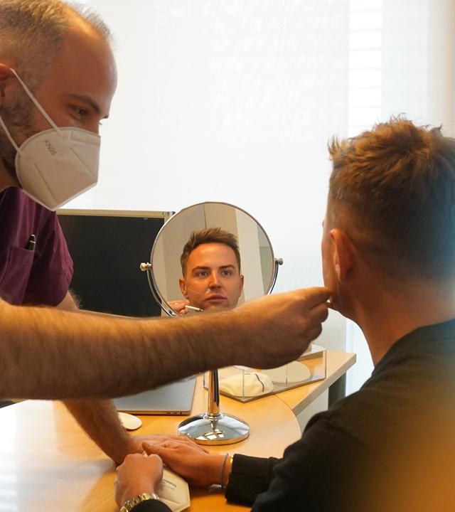 Zwei Personen sind in einem Raum zu sehen. Rechts sitzt ein Mann an einem Tisch, mit dem Rücken zur Kamera. Er blickt in einen Tischspiegel, sein Gesicht ist im Spiegelbild erkennbar. Links von ihm sitzt ein Mann mit Gesichtsmaske. Mit der rechten Hand hält er einen weißen Stift an das Gesicht des jungen Mannes.