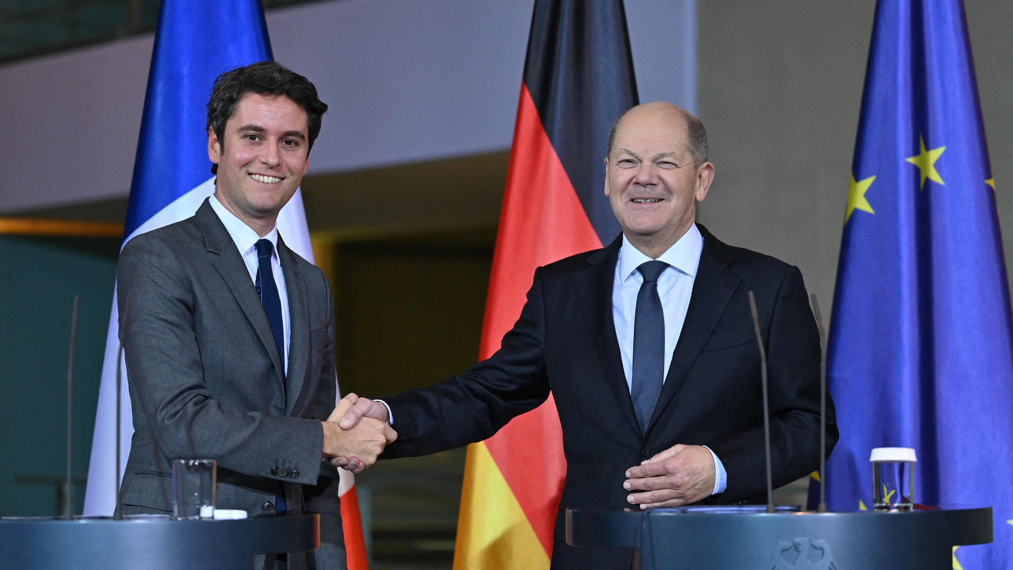 Bundeskanzler Olaf Scholz und Frankreichs Premierminister Gabriel Attal beim Treffen in Berlin. Sie stehen hinter zwei Redepulten und schütteln lächelnd die Hände.