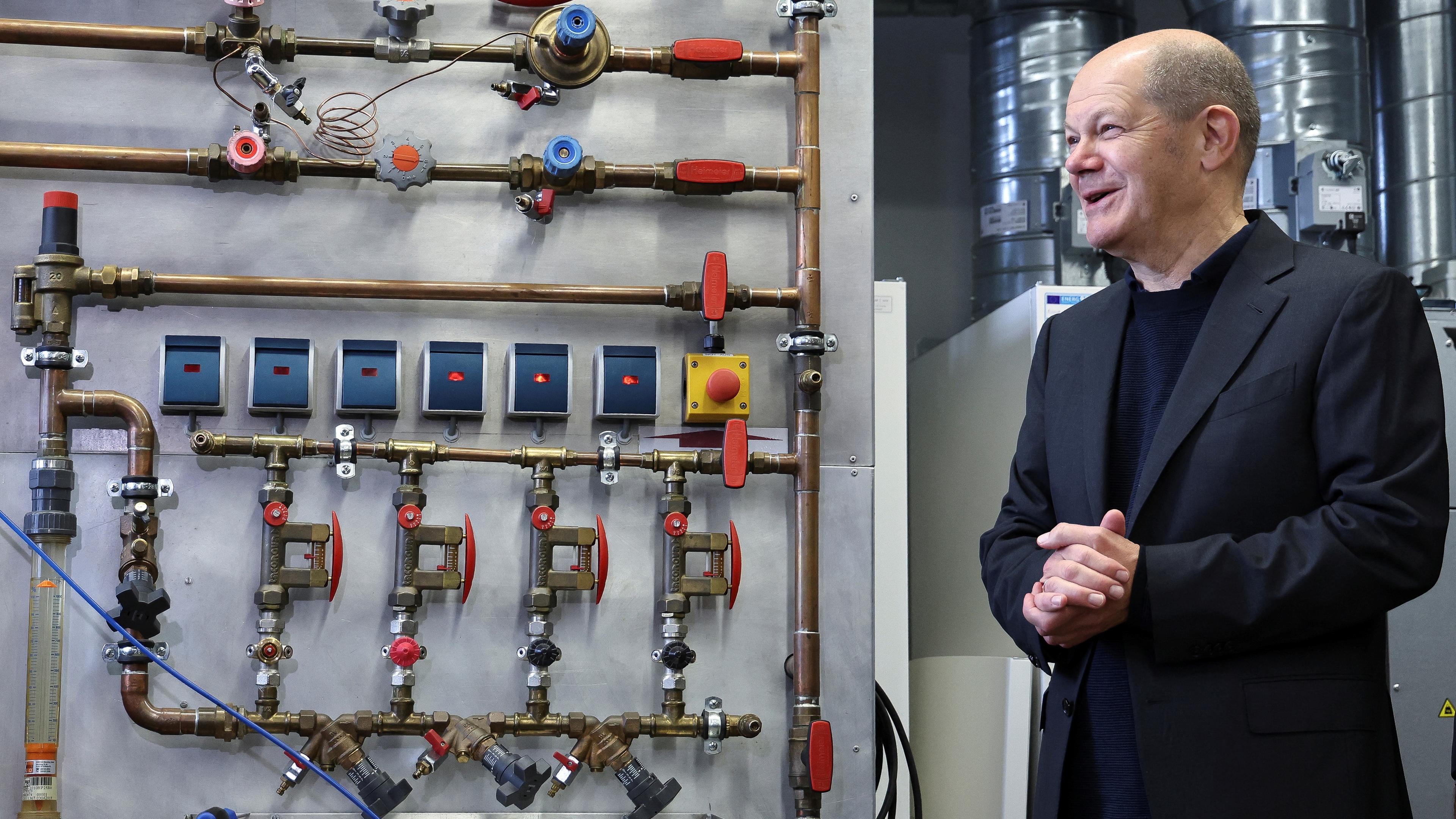 Scholz steht vor einer Hitzepumpe bei seinem Besuch von Mitarbeitenden des Energiesektors in München