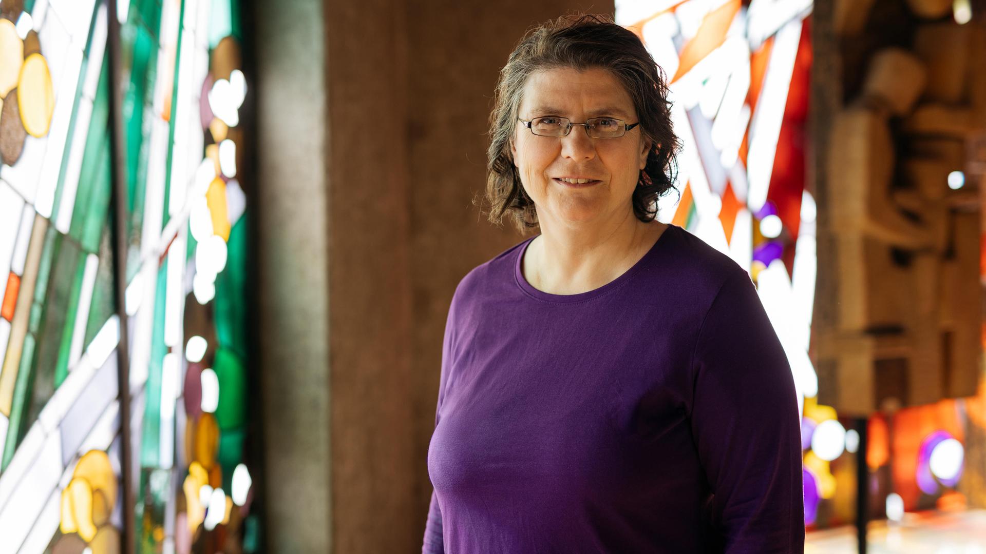 Christine Thürmer steht in einer Kirche, sie trägt ein lilafarbenes Oberteil und eine Brille und lächelt in die Kamera. Im Hintergrund sind bunte Kirchenfenster zu sehen. 