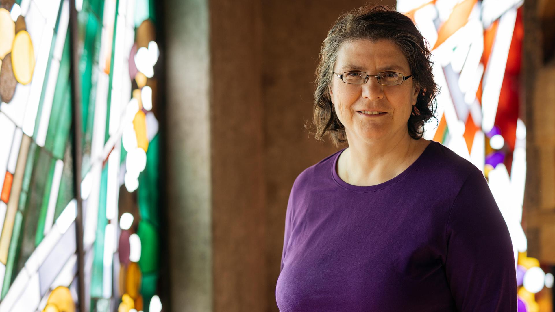 Christine Thürmer steht in einer Kirche, sie trägt ein lilafarbenes Oberteil und eine Brille und lächelt in die Kamera. Im Hintergrund sind bunte Kirchenfenster zu sehen. 