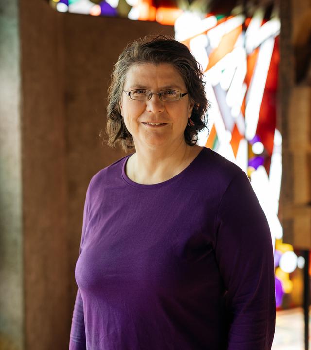 Christine Thürmer steht in einer Kirche, sie trägt ein lilafarbenes Oberteil und eine Brille und lächelt in die Kamera. Im Hintergrund sind bunte Kirchenfenster zu sehen. 