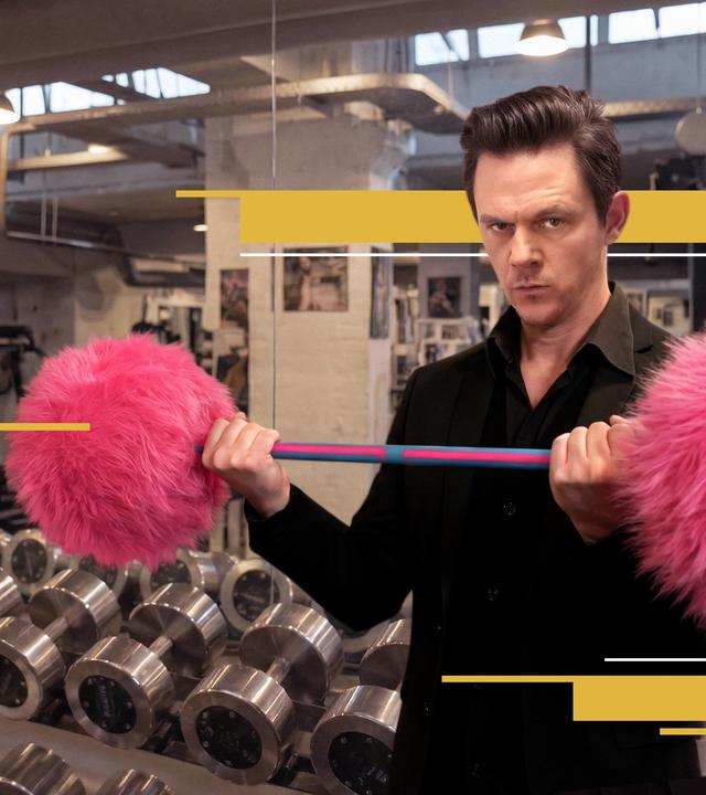 Moderator Jo Schück im  Fitnessstudio, umgeben von aufgereihten Kurzhanteln aus Metall. Er stemmt eine Hantel mit pinken Plüschgewichten. 