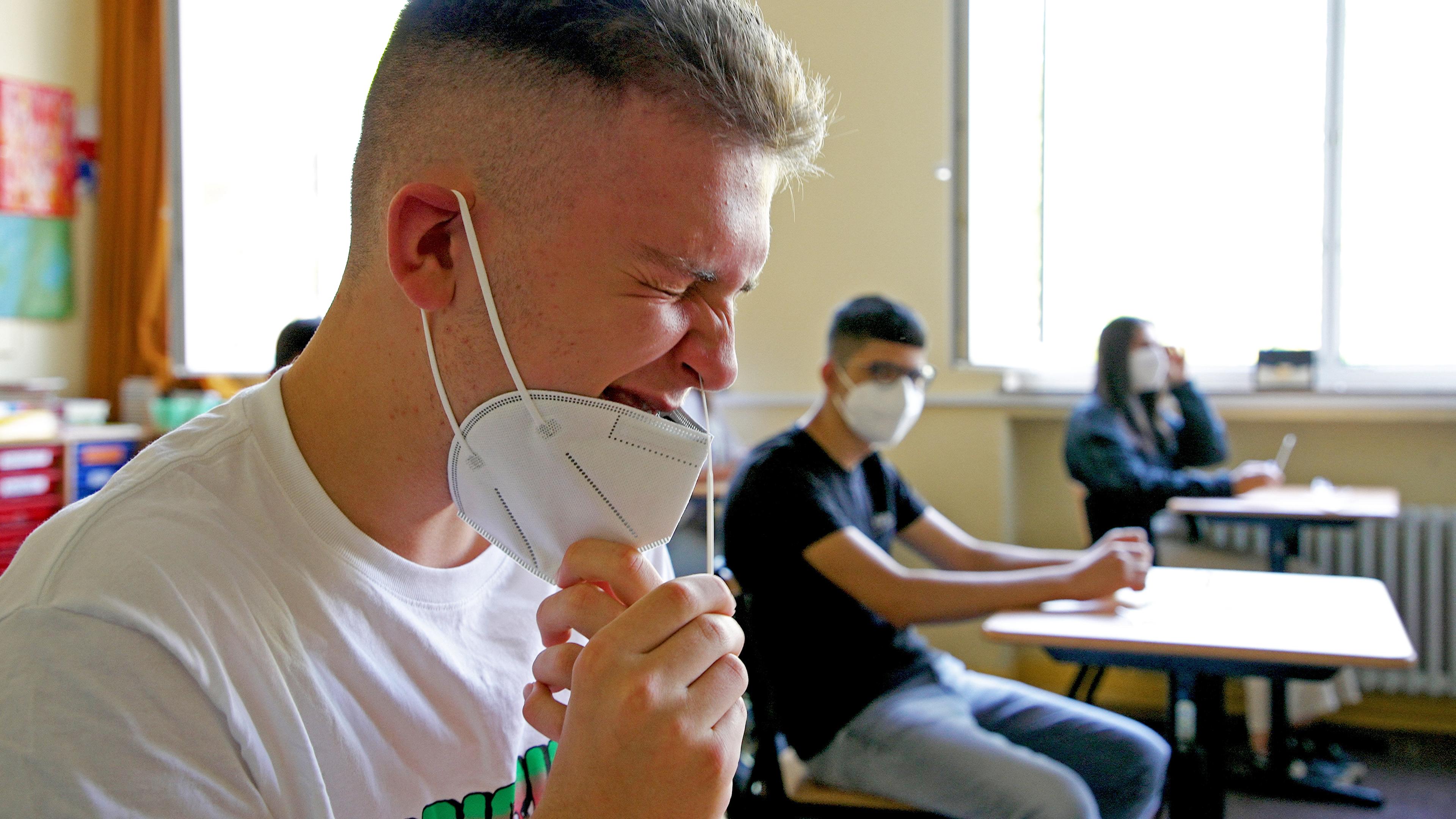 Ein Jugendlicher mit heruntergezogenem Mund-Nasenschutz und zusammengekniffenen Augen führt ein Teststäbchen in seine Nase ein.