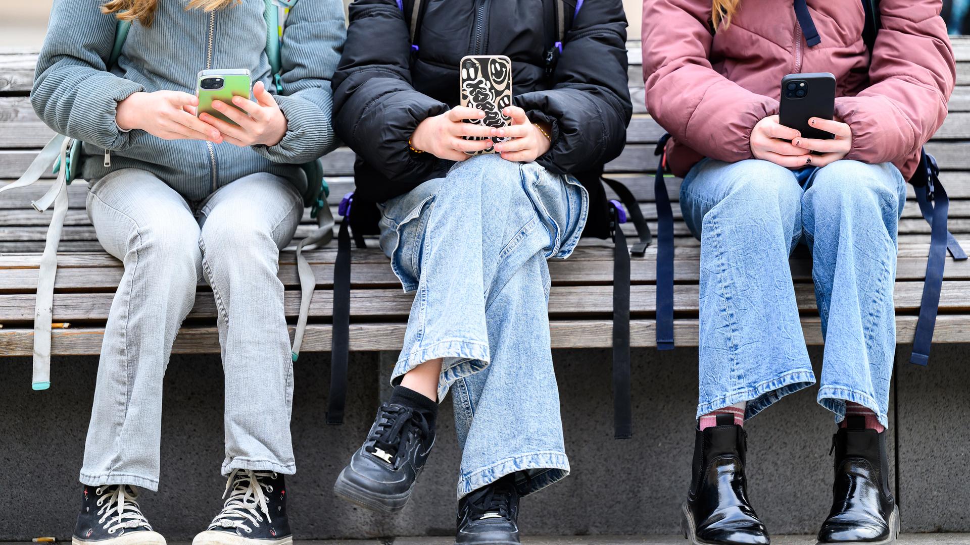 Drei Schülerinnen mit Schulranzen auf dem Rücken sitzen auf einer Parkbank und schauen auf ihr Handy. Die Köpfe sind auf dem Foto nicht zu sehen.