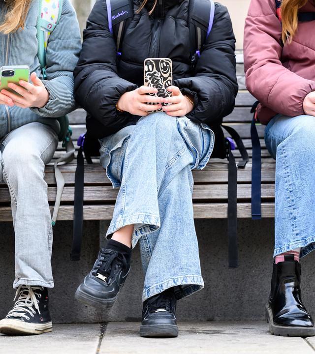 Drei Schülerinnen mit Schulranzen auf dem Rücken sitzen auf einer Parkbank und schauen auf ihr Handy. Die Köpfe sind auf dem Foto nicht zu sehen.