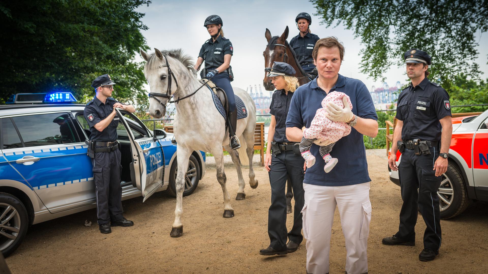 "Notruf Hafenkante - Schütteltrauma": Dr. Haase (Fabian Harloff) hält einen Säugling behutsam vor sich. Hinter ihm stehen die Polizeibeamten Melanie Hansen (Sanna Englund) und Mattes Seeler (Matthias Schloo). Außerdem im Bild zwei berittene Polizistinnen, ein Notarztwagen und ein Polizist, der an der Tür seines Dienstfahrzeuges steht.
