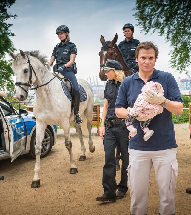 "Notruf Hafenkante - Schütteltrauma": Dr. Haase (Fabian Harloff) hält einen Säugling behutsam vor sich. Hinter ihm stehen die Polizeibeamten Melanie Hansen (Sanna Englund) und Mattes Seeler (Matthias Schloo). Außerdem im Bild zwei berittene Polizistinnen, ein Notarztwagen und ein Polizist, der an der Tür seines Dienstfahrzeuges steht.