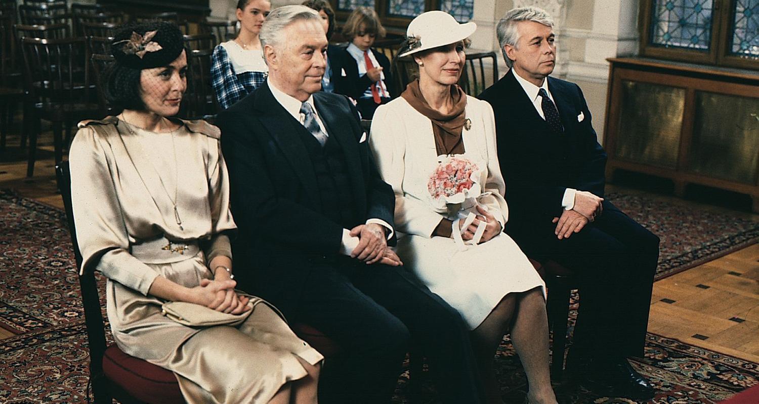 Werner und Angi sitzen in Abendgarderobe mit Onkel Rudolf und seiner Braut in einer Stuhlreihe im Saal des Standesamts. Im Hintergund sitzen die drei Kinder. 