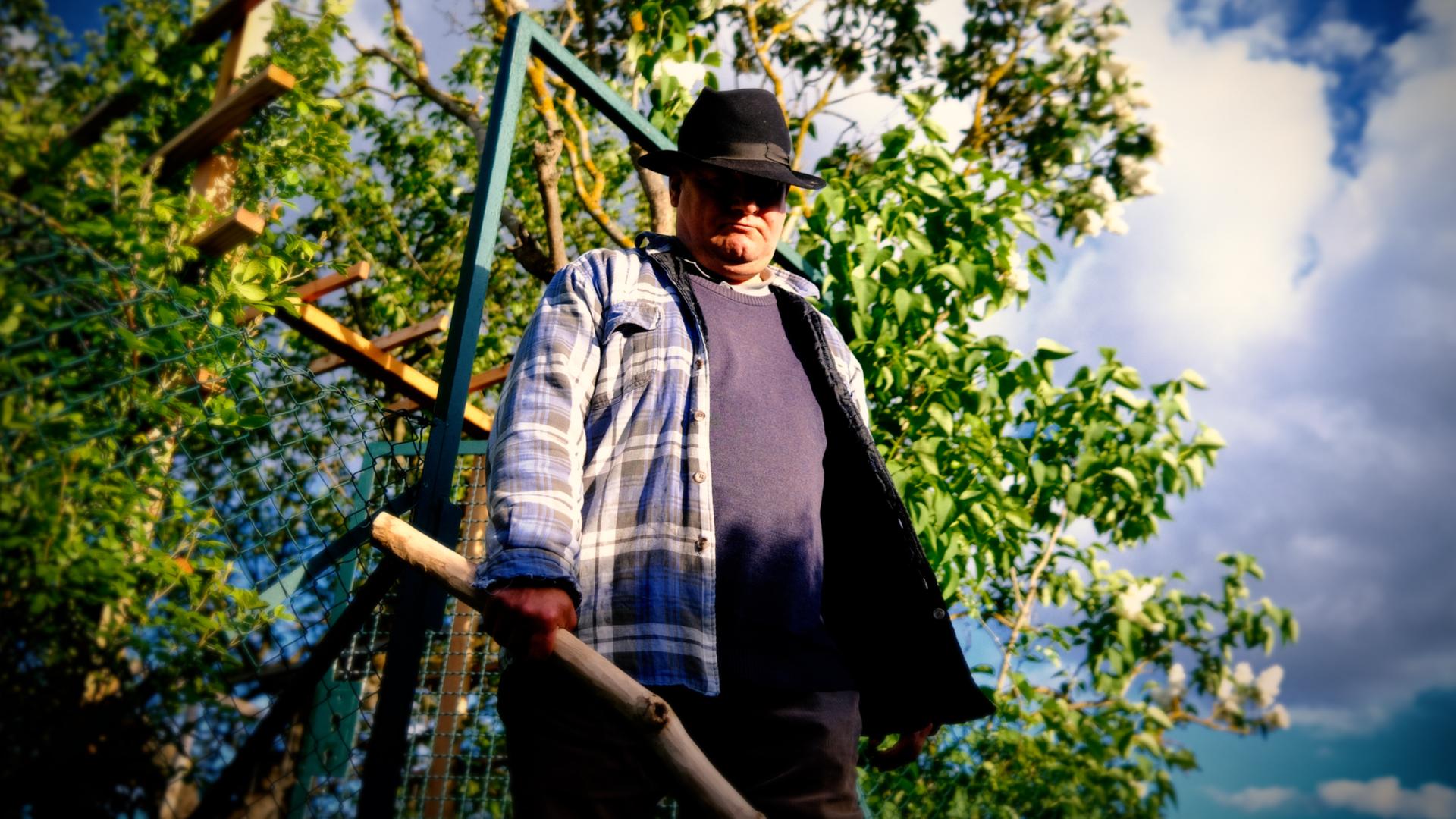 Ein Mann steht vor einem Gartentor und hält einen selbst geschnitzten Holzknüppel in der Hand.