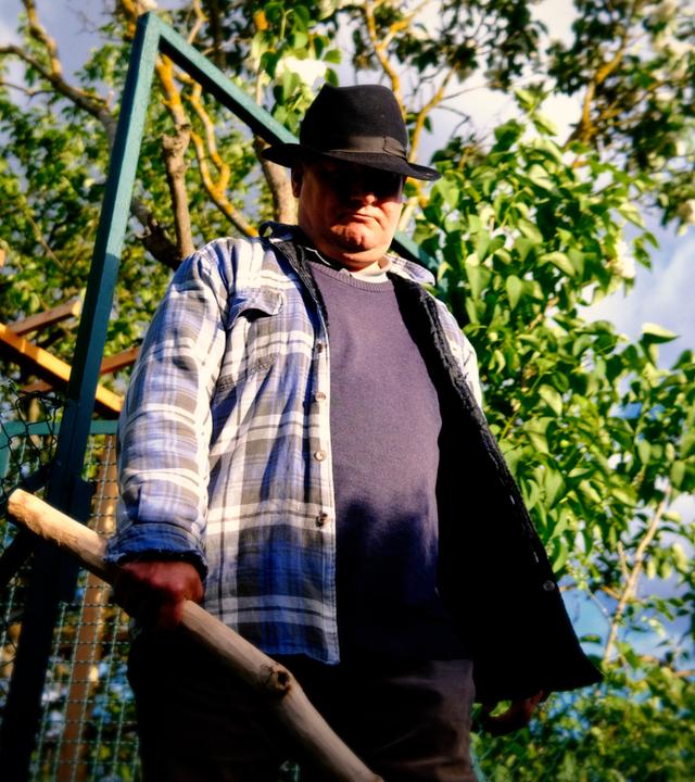 Ein Mann steht vor einem Gartentor und hält einen selbst geschnitzten Holzknüppel in der Hand.