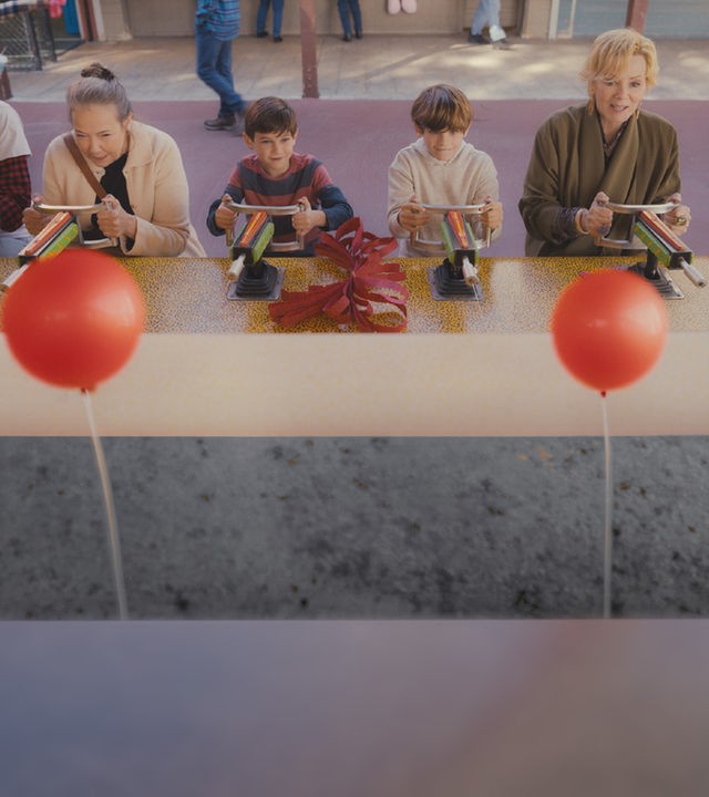 "Hacks - Schuld wirft Schatten": Deborah (Jean Smart), Susan (Harriet Sansom Harris) und deren beiden Enkel Mason (Henry Samiri) und Keller (Elaye Handal) stehen an einem Jahrmarktsstand und zielen konzentriert mit Wasserspritzpistolen. Im Vordergrund hängen zwei rote Luftballons.