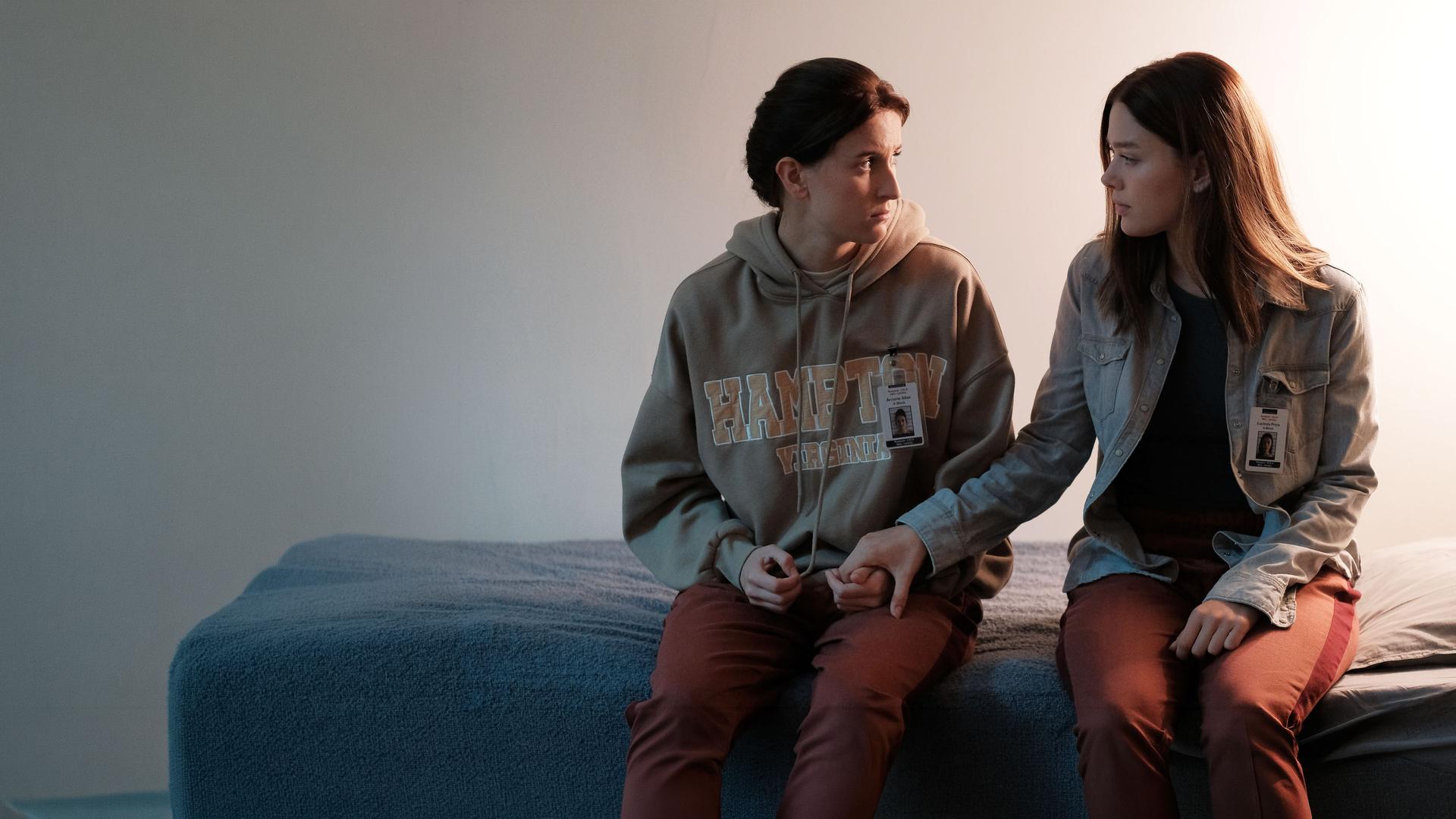 "Fallen - Schuldgefühle": Arriane (Josefine Koenig) und Luce (Jessica Alexander) sitzen nebeneinander auf einem Bett und halten sich an den Händen. Beide blicken sich dabei ruhig und verständnisvoll an.