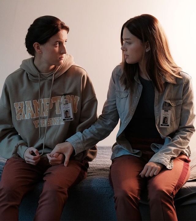 "Fallen - Schuldgefühle": Arriane (Josefine Koenig) und Luce (Jessica Alexander) sitzen nebeneinander auf einem Bett und halten sich an den Händen. Beide blicken sich dabei ruhig und verständnisvoll an.