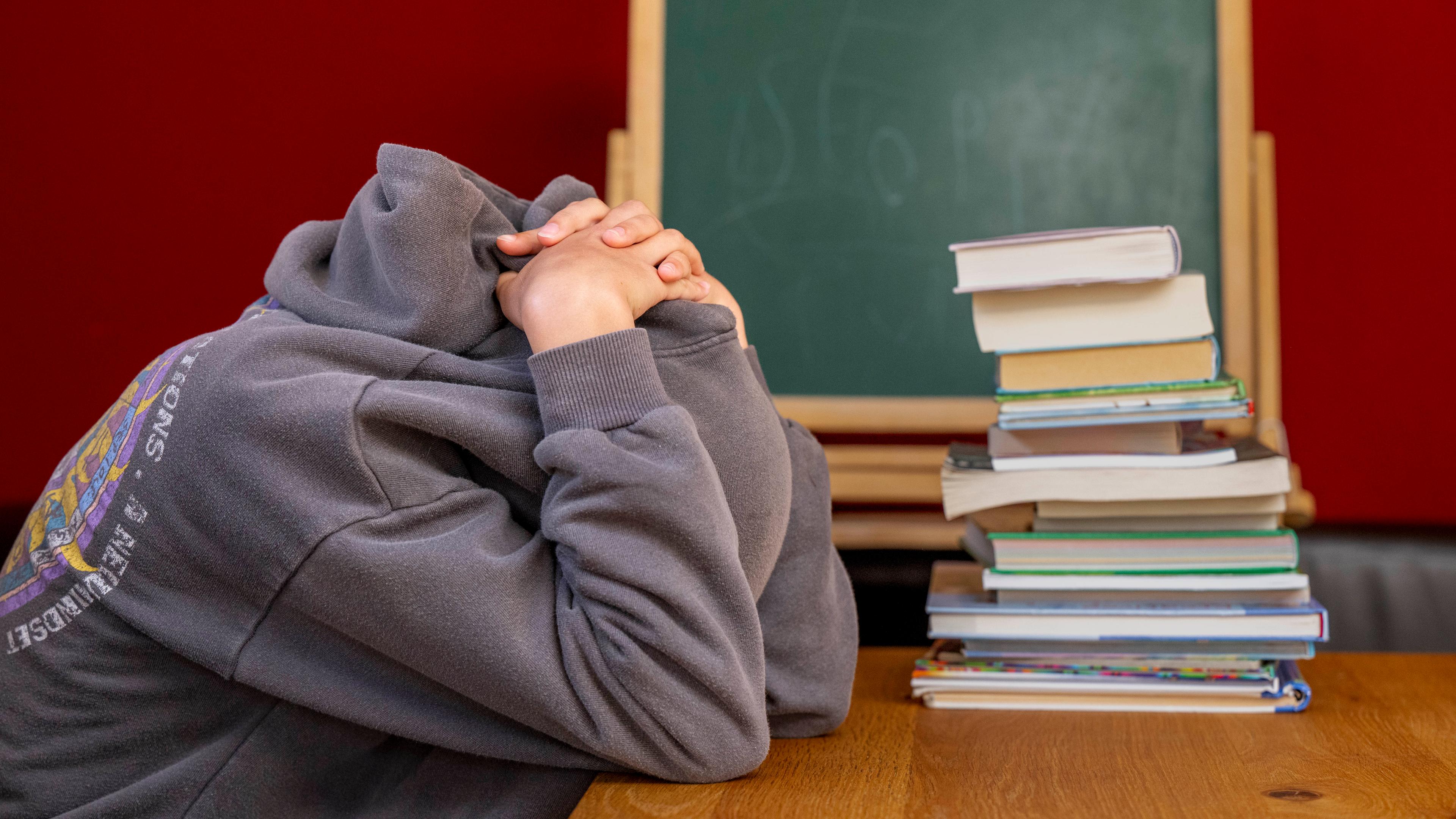 Eine Schülerin sitzt vor einem Stapel Büchern und hat trotzig die Kapuze ihres Pullovers über den Kopf gezogen.