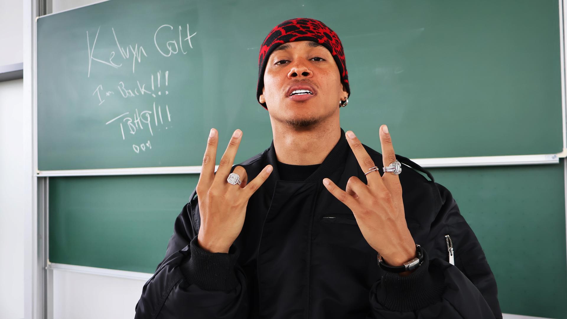 : Ein junger Mann steht vor einer grünen Schultafel, auf der "Kelvyn Colt" geschrieben ist, zusammen mit den Worten "I'm Back!!!". Der Mann trägt eine schwarze Jacke und eine rote, gemusterte Mütze. Er zeigt mit beiden Händen ein Handzeichen und trägt mehrere auffällige Ringe. Sein Gesichtsausdruck ist selbstbewusst, und er wirkt lässig und cool.