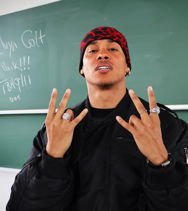 : Ein junger Mann steht vor einer grünen Schultafel, auf der "Kelvyn Colt" geschrieben ist, zusammen mit den Worten "I'm Back!!!". Der Mann trägt eine schwarze Jacke und eine rote, gemusterte Mütze. Er zeigt mit beiden Händen ein Handzeichen und trägt mehrere auffällige Ringe. Sein Gesichtsausdruck ist selbstbewusst, und er wirkt lässig und cool.