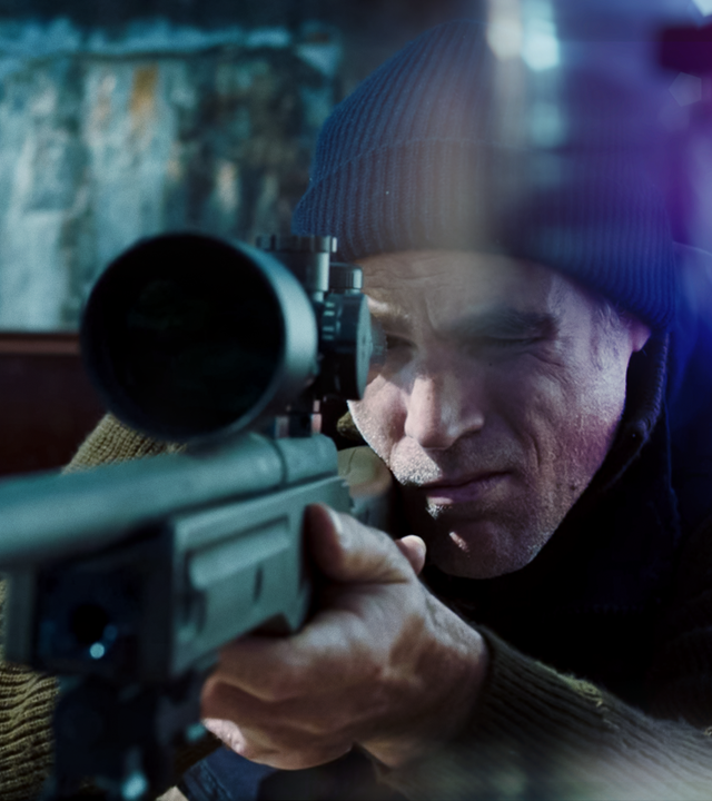 Ein Mann steht im Dunklen vor einem Fenster und setzt zum Schuss mit seiner Waffe an.