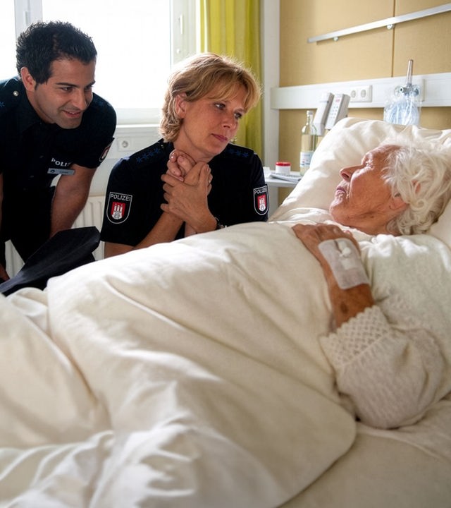 "Notruf Hafenkante - Schutzengel": Elsa (Renate Delfs) liegt in einem Krankenhausbett. Claudia (Janette Rauch), die neben ihr sitzt, hält liebevoll ihre Hand und sieht Elsa an. Tarik (Serhat Cokgezen), der ebenfalls neben dem Bett steht, lächelt die alte Dame aufmunternd an.
