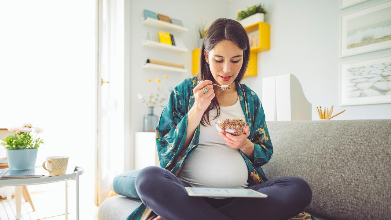 A pregnant woman sits on a sofa and eats a muesli.