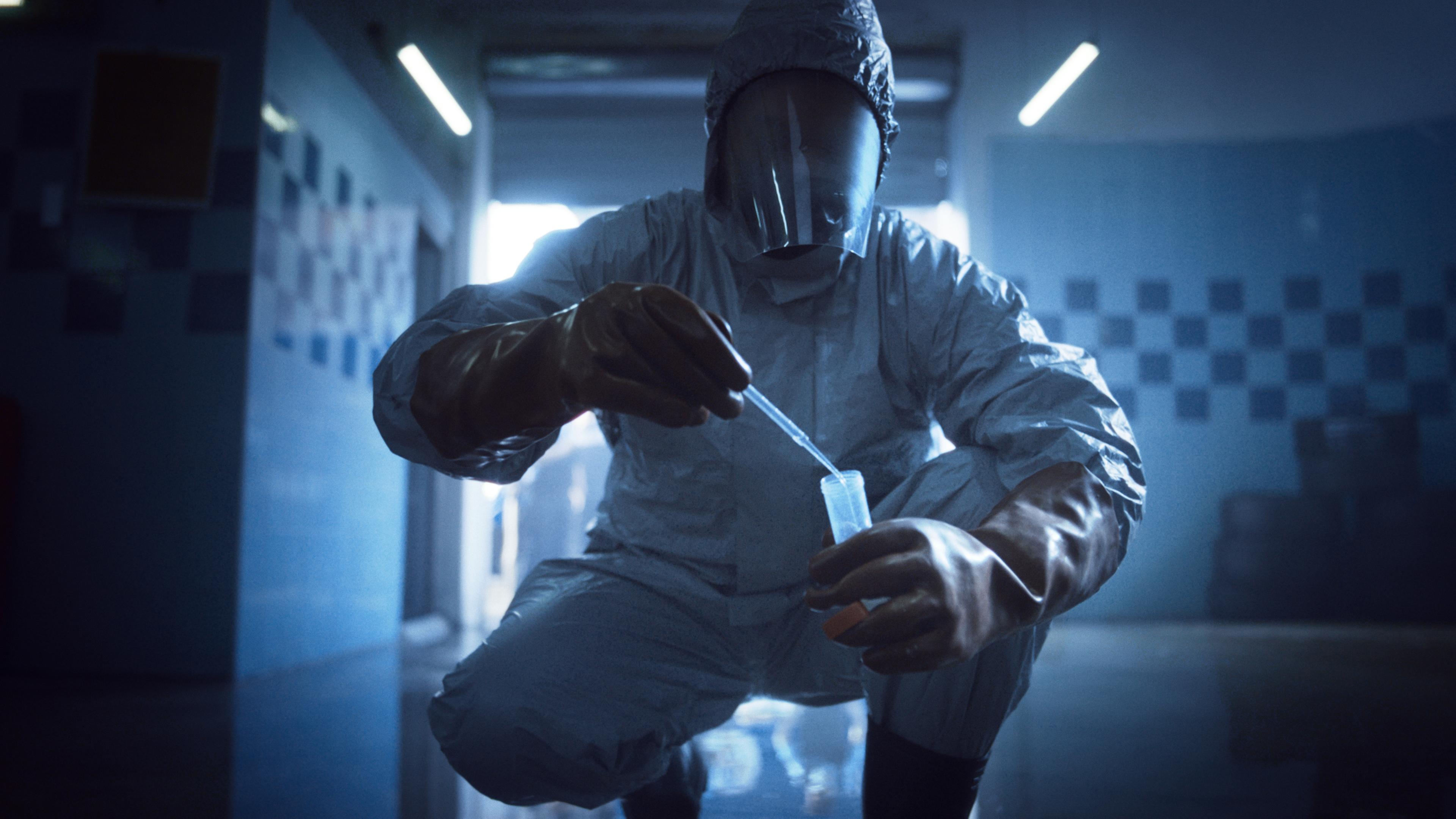 In einem gekachelten Raum sammelt jemand in einem Infektionsschutzanzug etwas mit einer Pipette in ein Glasröhrchen. 