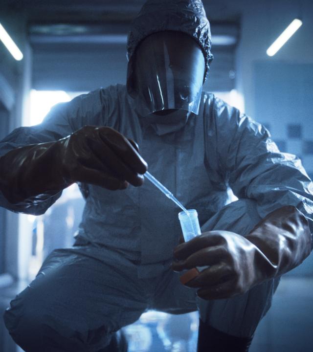 In einem gekachelten Raum sammelt jemand in einem Infektionsschutzanzug etwas mit einer Pipette in ein Glasröhrchen. 