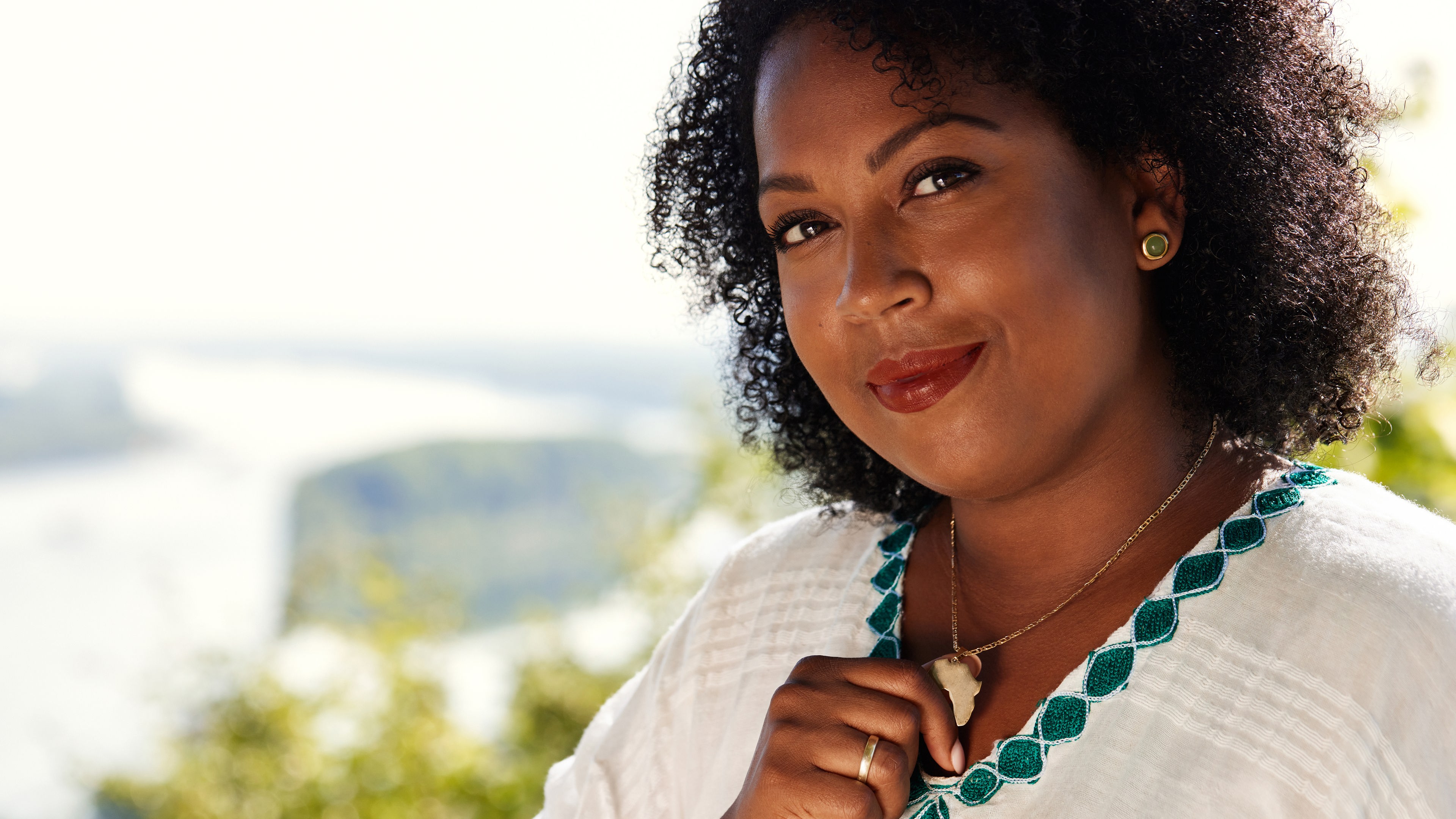 Menna Mulugeta steht vor einem Landschaftspanorama des Rheins. Sie fasst mit einer Hand an einen Kettenanhänger in Form von Afrika.