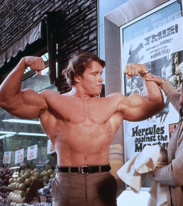 Ein Bodybuilder posiert mit nacktem Oberkörper, während eine Frau ihn am Arm fasst.