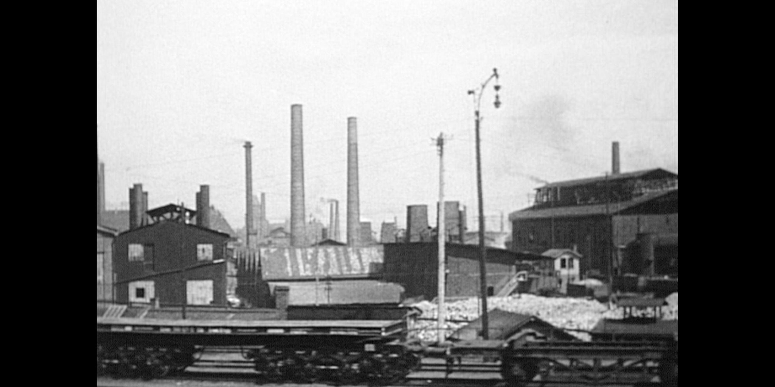 schwarz-weiß Bild, im Vordergrund Bahngleise, im Hintergrund mehrere Schornsteine und Strommasten. 