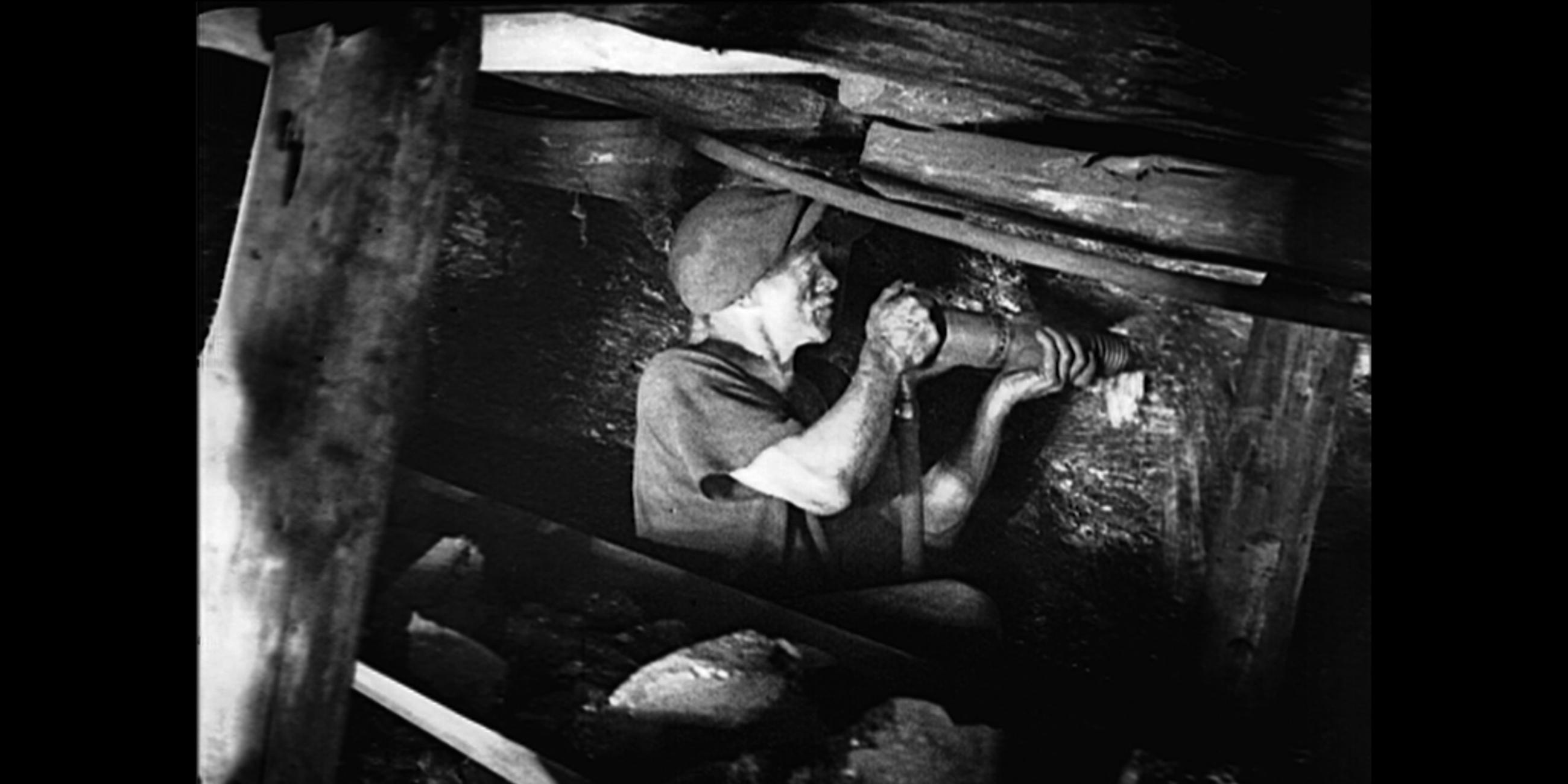 Schwarz-weiß Fotografie eines Bergarbeiters im Stollen, der bohrt.