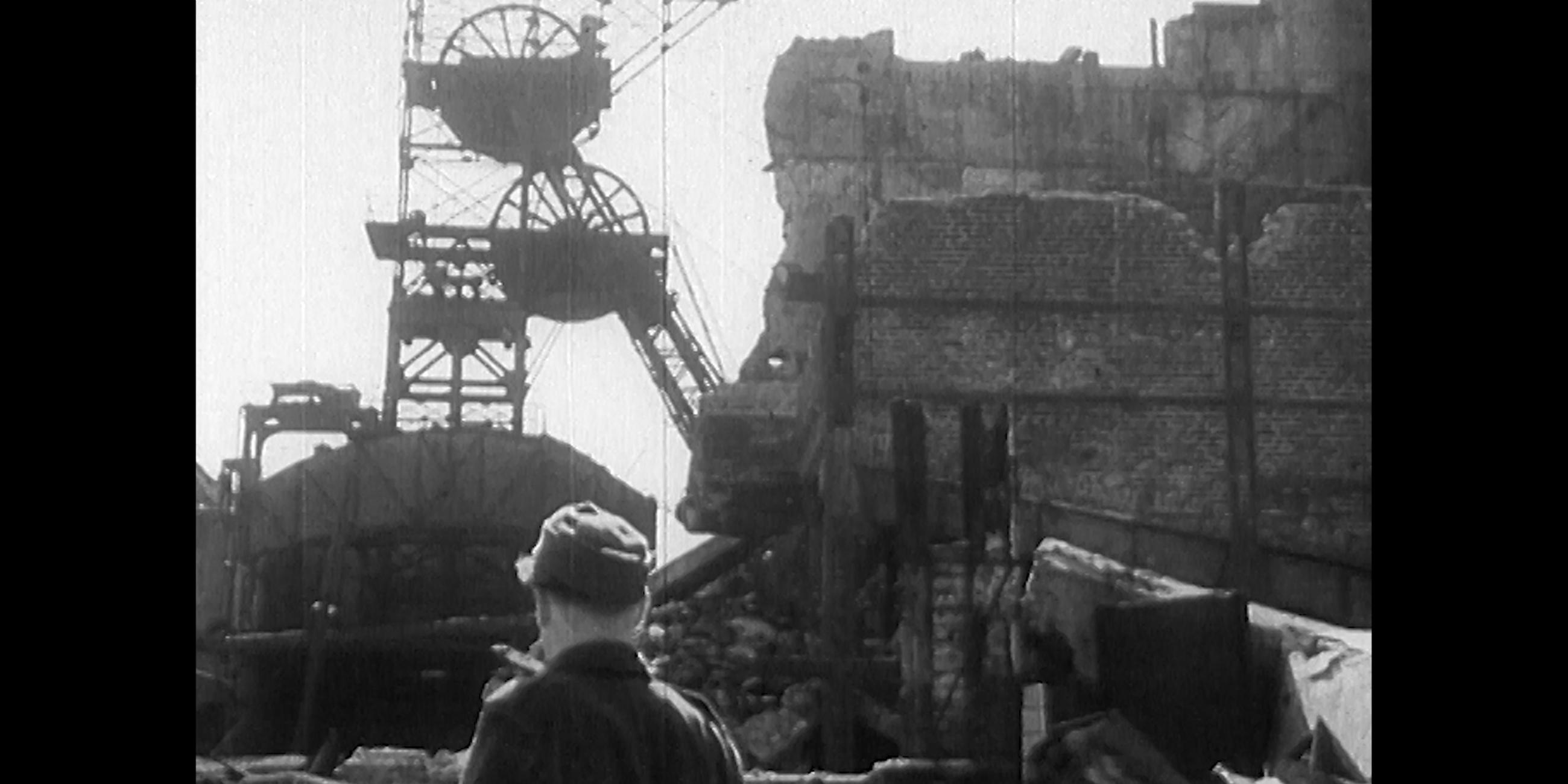 Schwarz-weiß Fotografie eines Bergarbeiters, der vor einem großen Trümmerhaufen aus Stein steht. 