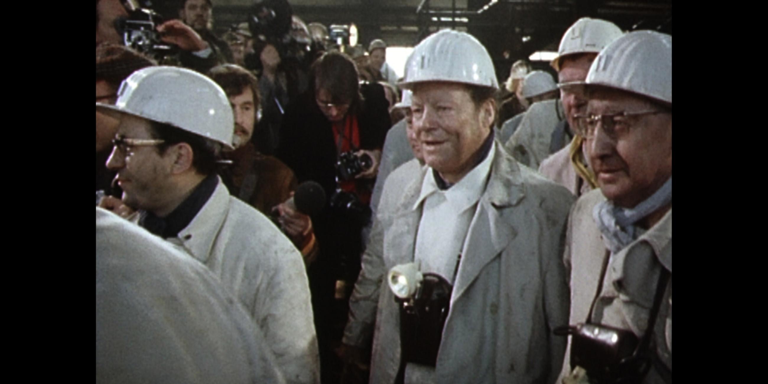 Willy Brandt im Schutzkittel umringt von Bergarbeitern mit Helmen und Journalisten im Hintergrund. 