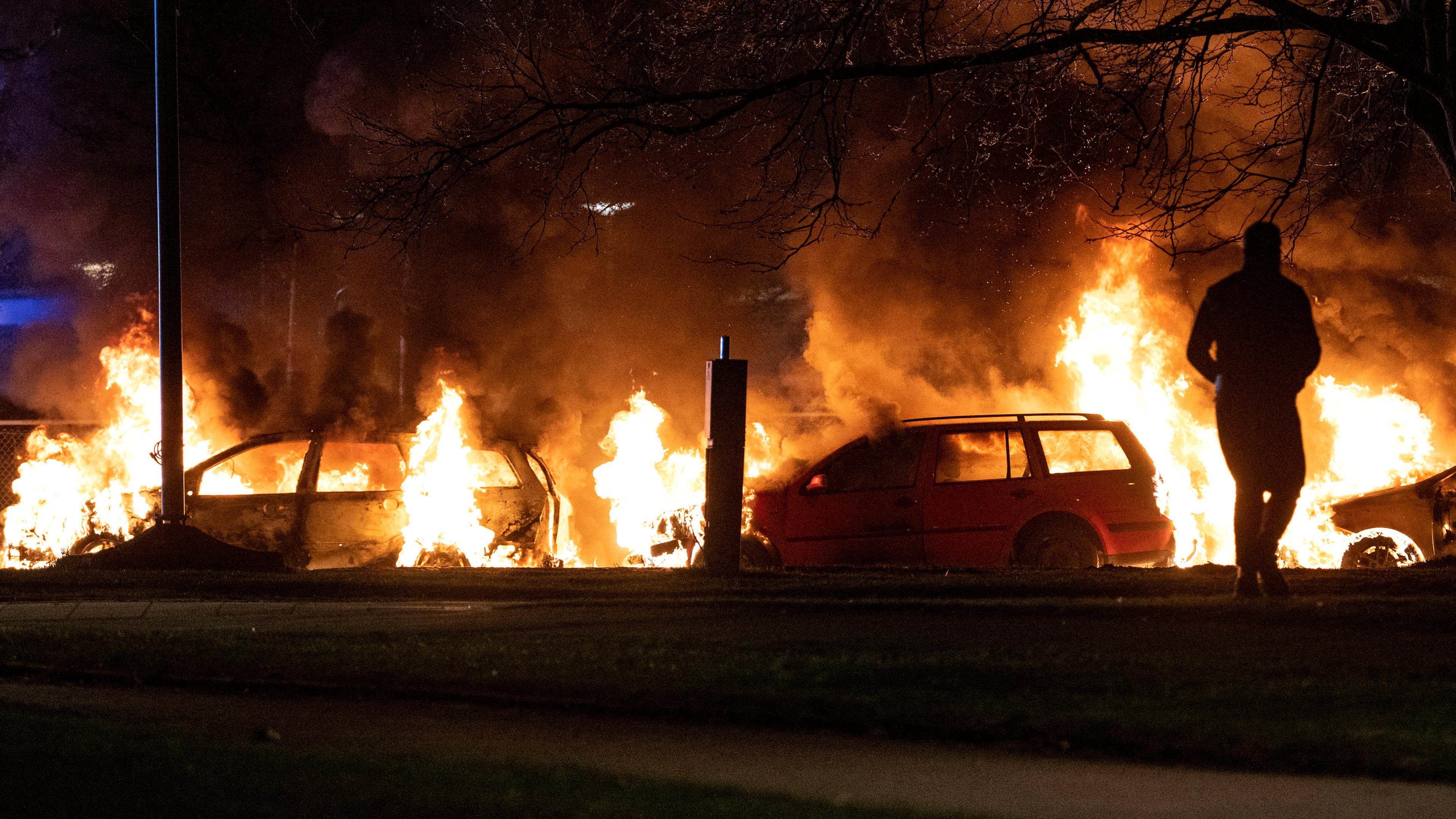 Autos in Flammen, davor Silhouette eines Menschen