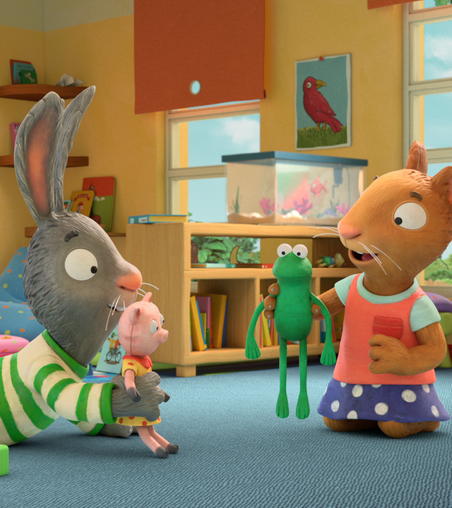 Pip liegt auf dem Bauch und hält Schweinchen in der Hand. Posy kniet und hebt Froschi hoch. Die beiden spielen mit den Kuscheltieren. Im Hintergrund sitzen Frankie und Jamila auf Sitzsäcken und lesen.