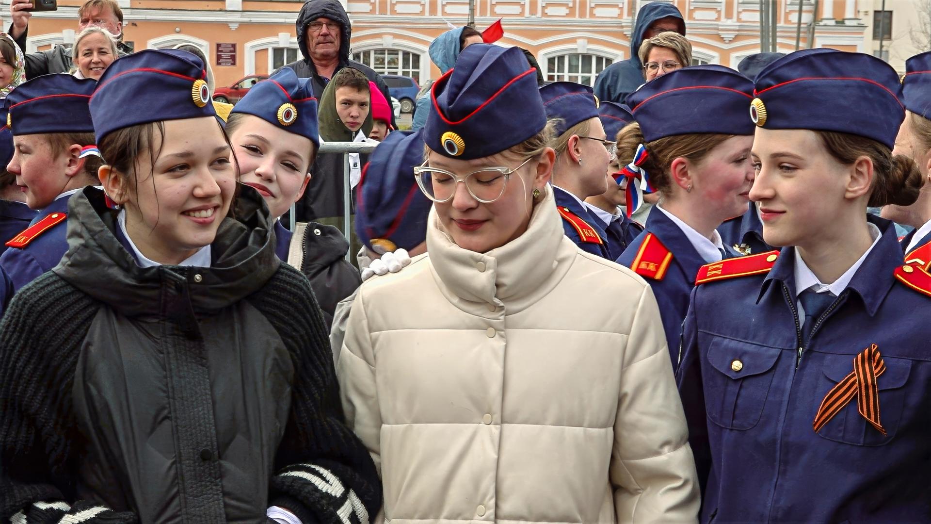 Eine Gruppe junger Menschen in blauen Uniformen mit Mützen und rot-goldenen Abzeichen, einige mit schwarzer Schleife, stehen lächelnd zusammen bei einer öffentlichen Veranstaltung.