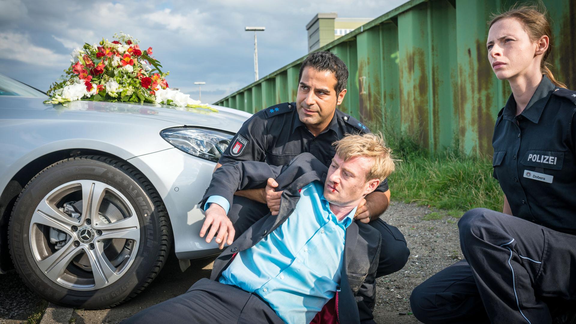 "Notruf Hafenkante - Schwiegermuttermonster": Die Polizisten Tarik Coban (Serhat Cokgezen) und Kim Dobers (Carolyn Genzkow) sitzen in der Hocke vor einem mit einem Brautbouquet geschmückten silbernen Wagen. Tarik stützt Theo Metzner (Eric Bouwer), der auf dem Boden liegt und aus der Nase blutet.