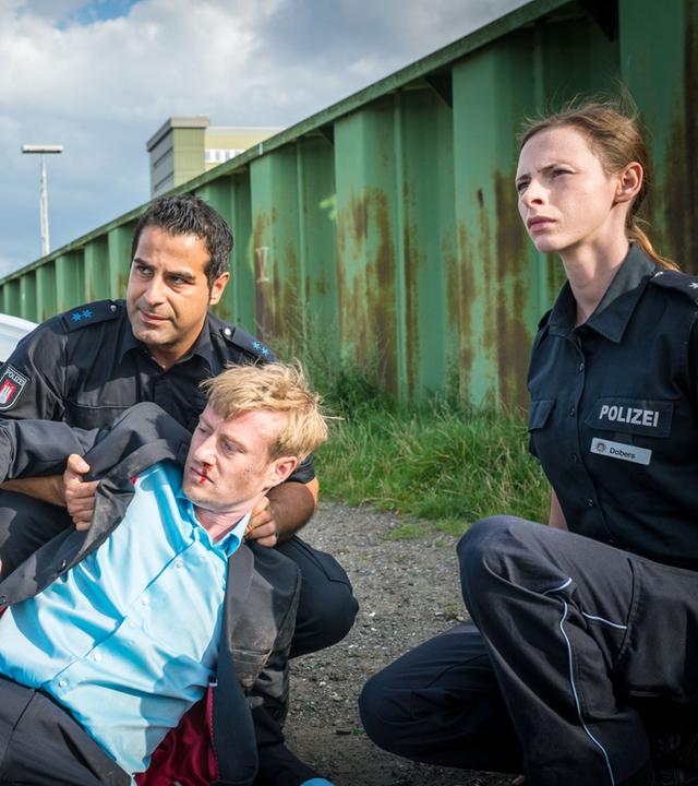 "Notruf Hafenkante - Schwiegermuttermonster": Die Polizisten Tarik Coban (Serhat Cokgezen) und Kim Dobers (Carolyn Genzkow) sitzen in der Hocke vor einem mit einem Brautbouquet geschmückten silbernen Wagen. Tarik stützt Theo Metzner (Eric Bouwer), der auf dem Boden liegt und aus der Nase blutet.