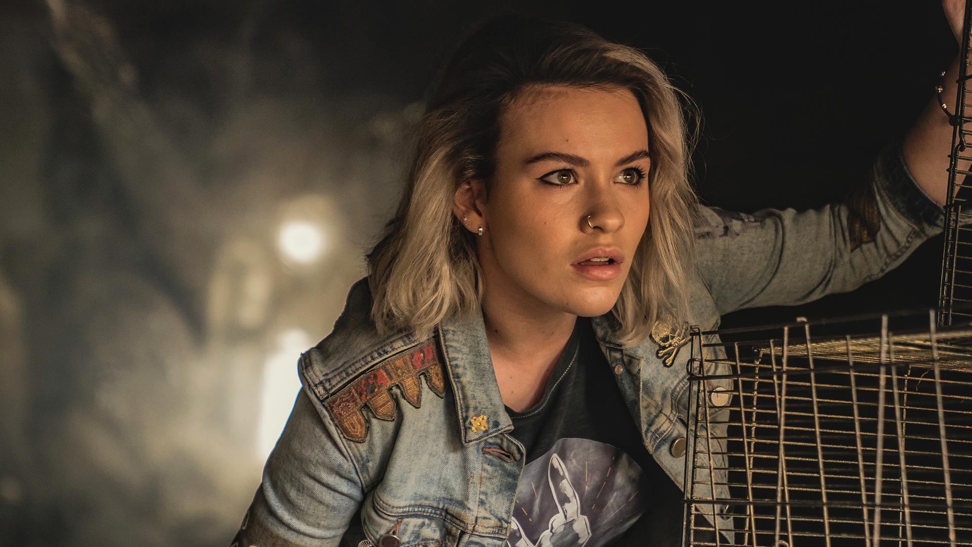 Issie trägt eine Jeansjacke und eine karierte Hose. Sie kniet neben Käfigen, in denen Stroh liegt, und beobachtet etwas. Der Hintergrund ist dunkel.