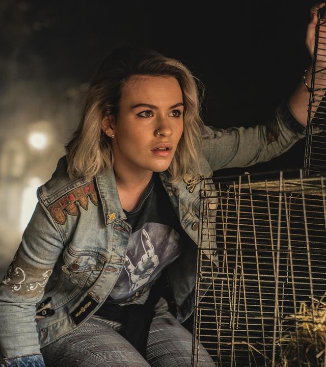 Issie trägt eine Jeansjacke und eine karierte Hose. Sie kniet neben Käfigen, in denen Stroh liegt, und beobachtet etwas. Der Hintergrund ist dunkel.