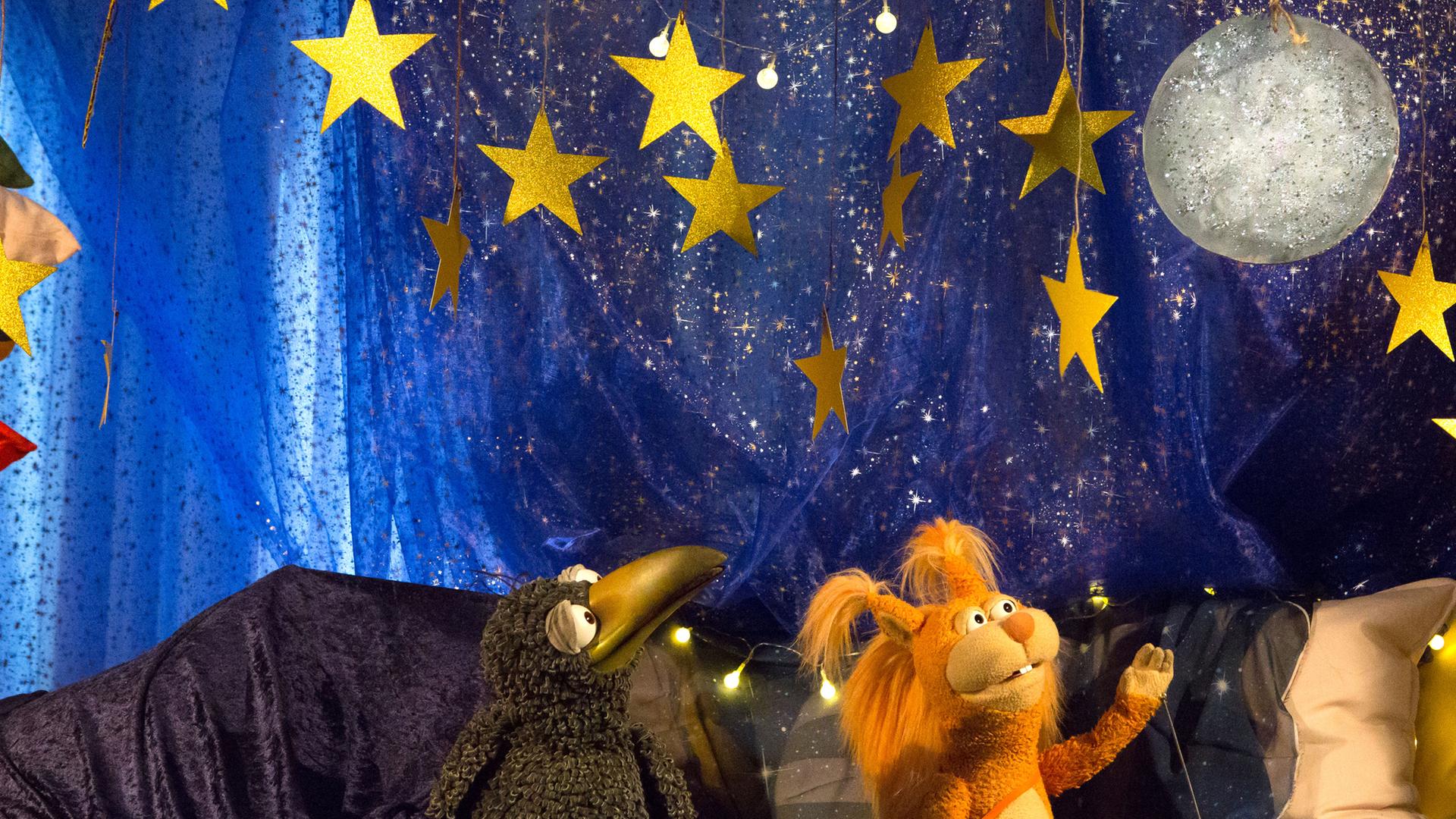 Rudi und Ella hocken in der Kuschelecke, die nächtlich dekoriert ist. Beide schauen fasziniert auf einen Glitzermond, der über ihnen am Sternenhimmel hängt. Ella hebt einen Arm in seine Richtung. 