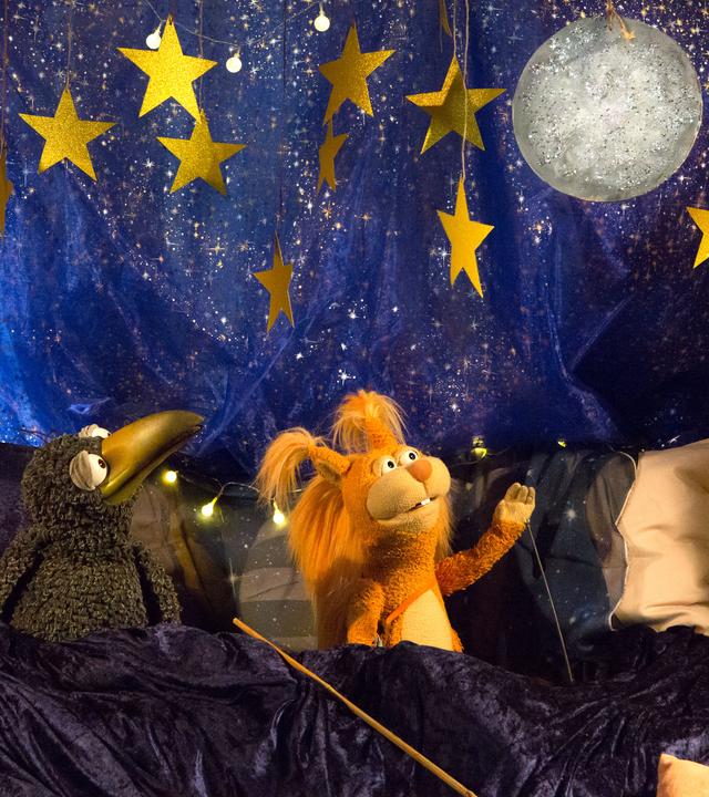 Rudi und Ella hocken in der Kuschelecke, die nächtlich dekoriert ist. Beide schauen fasziniert auf einen Glitzermond, der über ihnen am Sternenhimmel hängt. Ella hebt einen Arm in seine Richtung. 