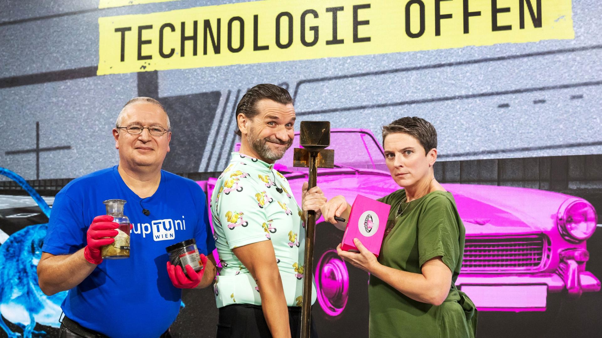 Das Bild zeigt drei Personen, die nebeneinander stehen. Im Hintergrund ist ein grafisches Muster mit der Aufschrift "IHR HABT WOHL DIE TECHNOLOGIE OFFEN" in gelben Buchstaben auf einem dunkelgrauen Untergrund. Die erste Person links trägt ein blaues T-Shirt mit der Aufschrift "upTuni Wien" und hält eine Glasflasche sowie ein schwarzes Behältnis in den Händen. Sie hat eine Brille auf und trägt rote Handschuhe.   In der Mitte steht eine zweite Person in einem hellen Hemd mit einem bunten Muster aus kleinen Figuren. Diese Person hat eine verspielte Miene und hält einen langen Stock.   Die dritte Person auf der rechten Seite trägt ein grünes Oberteil und sieht ernst aus. Sie hält ein pinkes Notizbuch oder ein ähnliches Objekt in der Hand und hat eine kurze Frisur. Im Hintergrund sind farbenfrohe grafische Elemente, darunter ein pinkes Auto und eine cartoonartige Illustration eines Tieres. Die gesamte Szenerie hat einen modernen und dynamischen Charakter.